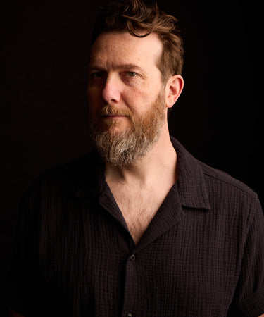 John Mark McMillan wearing a dark textured short-sleeve button-up shirt, photographed against a solid black background.